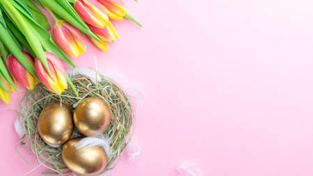 Easter Golden eggs in basket with spring tulips, white feathers on pastel pink background in Happy Easter decoration. Spring holiday flat lay conceptの写真素材