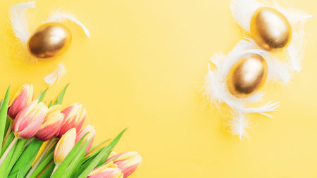 Easter symbol. Golden eggs in basket with spring tulips, white feathers on pastel yellow background in Happy Easter decoration. Congratulatory easter flat lay designの写真素材