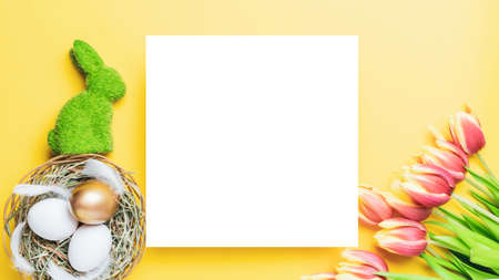 Easter eggs background. Golden, white color egg in basket with spring tulips, feathers on pastel yellow background in Happy Easter decoration. Foil minimalist egg design, modern top view templateの写真素材