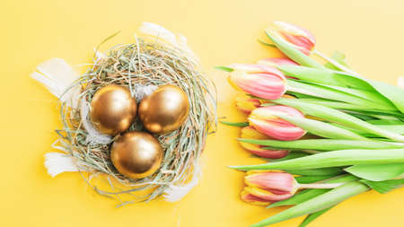 Easter Golden eggs in basket with spring tulips, white feathers on pastel yellow background in Happy Easter decoration. Spring holiday flat lay conceptの写真素材