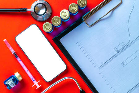 Medical equipment. Doctor stethoscope, healthcare charts, syringe with needle and black smartphone with blank screen on hospital red background. For design application, website projectの写真素材