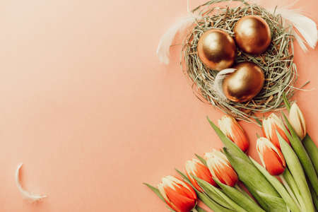 Egg color. Happy Easter decoration: golden color eggs in basket with spring tulips, white feathers on pastel pink background. Foil minimalist egg design, modern top view templateの写真素材