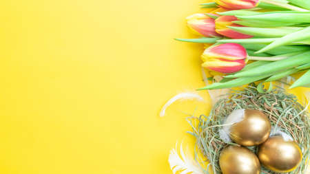 Egg color. Happy Easter decoration: golden color eggs in basket with spring tulips, white feathers on pastel yellow background. Foil minimalist egg design, modern top view templateの写真素材