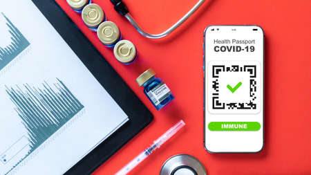 Immune passport. Coronavirus immunity certificate or vaccine passport on smartphone screen with doctor stethoscope, healthcare charts, syringe and medical equipment on hospital red backgroundの写真素材