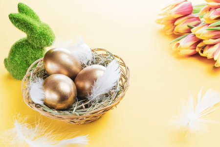Easter Golden eggs in basket with spring tulips, white feathers on pastel yellow background in Happy Easter decoration. Spring holiday conceptの写真素材
