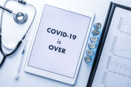 End of lockdown. Doctor stethoscope, hospital healthcare charts, syringe with needle and white tablet with text on screen on medical equipment background. COVID is over conceptの写真素材
