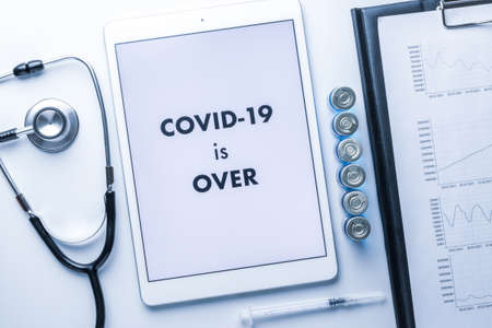 End of lockdown. Doctor stethoscope, hospital healthcare charts, syringe with needle and white tablet with text on screen on medical equipment background. COVID is over conceptの写真素材