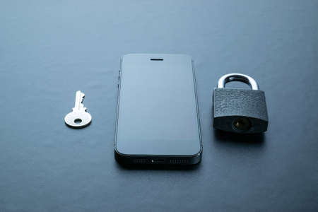 Technology security. Modern space grey mobile phone with padlock, key on dark background. Smartphone security breach concept.の写真素材