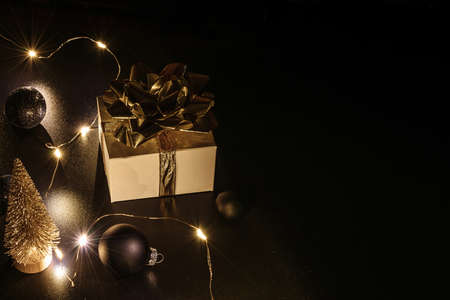 Gift boxes background. White gift box with golden ribbon, New Year balls and sparkling lights garland in Christmas composition on black for greeting card. Winter festive composition with copy spaceの写真素材