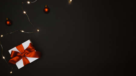 Holiday winter background. White gift box with red ribbon, New Year balls and sparkling lights garland in Christmas composition on black for greeting card. Flat lay, top view, copy space.の写真素材