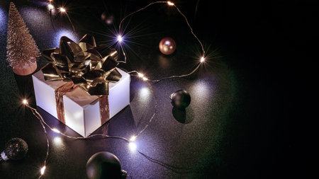 Surprise gift. White gift box with golden ribbon, New Year balls and sparkling lights garland in...