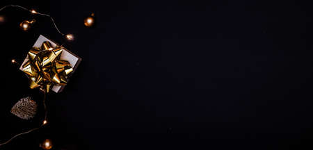 Holiday winter golden. White gift box with golden ribbon, New Year balls and sparkling lights garland in Christmas composition on black background for greeting card. Festive decoration, copy spaceの写真素材