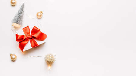 Winter background gold. White gift with red bow, gold balls and sparkling lights garland in xmas decoration on white background for greeting card. Winter festive composition with copy spaceの写真素材