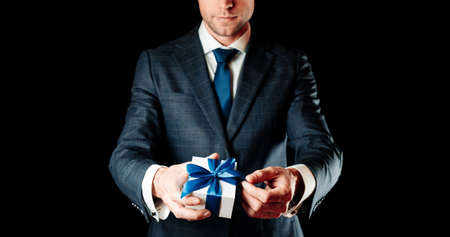 Holding gift box. Happy young business man holding surprise giftbox present with blue ribbon isolated on black background. Present for birthday, valentine day, Christmas, New Yearの写真素材