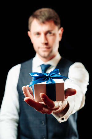 Gift in hand. Happy young business man holding surprise gift box present with blue ribbon isolated on black background. Black Friday, Valentine Day congratulations bannerの写真素材