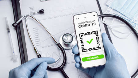 Covid passport doctor holding. Coronavirus vaccination certificate of immunity passport on smartphone screen with doctor stethoscope, syringe and medical equipment on hospital white backgroundの写真素材
