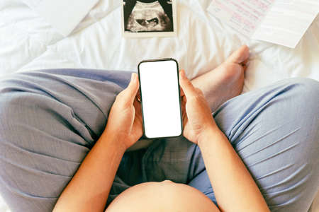 Pregnancy smartphone screen. Pregnant woman holding smartphone mock up. Mobile pregnancy online maternity application. Concept of pregnancy, maternity, expectation for baby birthの写真素材