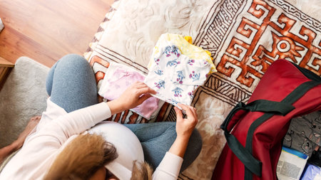 Pregnancy bag baby clothes. Happy young pregnant woman holding yellow baby clothes. Mother packing suitcase, bag for maternity hospital. Concept of pregnancy, maternity, expectation for baby birthの写真素材