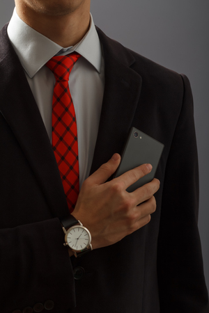 Young man in black suit keeping his hand with wristwatch on his chest and holding a phoneの写真素材
