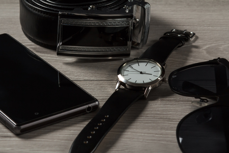 Watch with a leather strap, leather belt with metal buckle, sell phone, black sunglasses on a gray wooden backgroundの写真素材