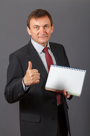 Confident and friendly elegant handsome mature businessman standing in front of a grey background holding a notebookの写真素材