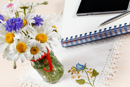 Daisy chamomile and cornflowers in vase on a wooden table with a smart-phone, exercise book and a penの写真素材
