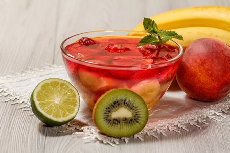 Cherry jelly with strawberry pieces in the glass bowl topped mint leaves, nectarine, kiwi, lime and banana on the white napkinの写真素材