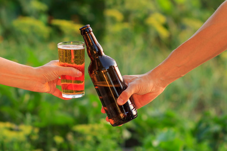 Cropped image of woman and man clanging glass of beer and bottle of beer together on natural green blurred background. Woman clinked her glass against man with bottleの写真素材