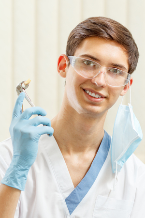Portrait of handsome male dentist with stainless steel dental tongs or pliers and extracted lower tooth in it. Doctor wearing white uniform, glasses and blue glovesの写真素材