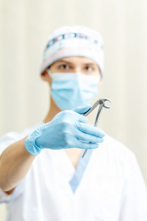 Portrait of handsome male dentist with stainless steel dental tongs or pliers for extraction of lower teeth in the dental clinic. Selective focus on dental tongsの写真素材