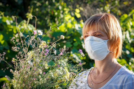 Unhappy woman in protection mask holding bouquet of wildflowers and trying to fight allergies to pollen. Woman protecting her nose from allergens. Allergy concept.の写真素材