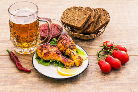 Grilled chicken legs roasted on the grill and lettuce leaves on plate with tomato sauce in a bowl, fresh tomatoes, bitter pepper and black bread with sunflower seeds, glass mug of beer. BBQ, picnicの写真素材