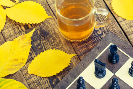 Chess pieces on board, cup of tea and autumn yellow leaves on wooden background