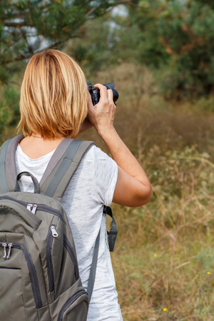 Young lady walking on at forest with digital camera. Hiker with backpack taking pictures of the summer landscape outdoors in nature. Healthy active lifestyleの写真素材
