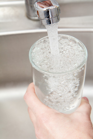 Glass in female hand is filled with clean transparent water under the tap with metal sink on the backgroundの写真素材