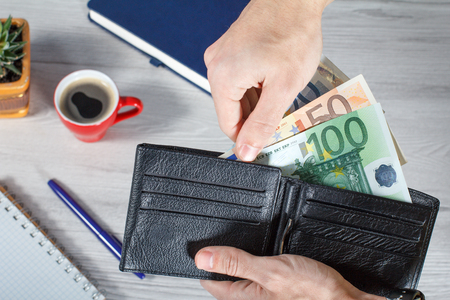 Male hands holding black leather wallet full of euro bills. Cup of coffee, notebook, pen and plant in a pot on background. Top viewの写真素材