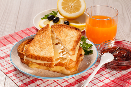 Toasted slices of bread with cheese and green parsley on white plate, glass og orange juice, lemon, olives, spoon and glass bowl with strawberry jam with red kitchen napkin. Good food for breakfastの写真素材