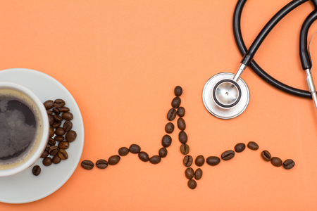 Part of cup of coffee on white saucer with coffee beans folded in the form of a cardiogram and phonendoscope on peach colored background. Top view. Health care conceptの写真素材