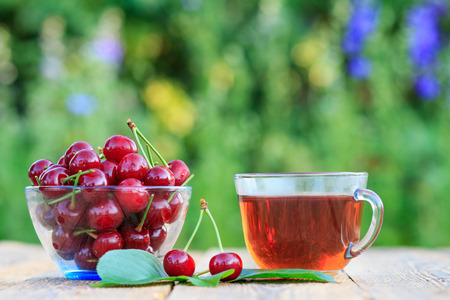 Red ripe cherry fruits with peduncles in glass bowl and cup of tea on old wooden boardsの写真素材