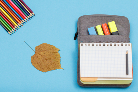 Color pencils, dry leaf, open exercise book on bag-pencil case with color felt pens and marker on blue background. Top view with copy space. Back to school concept. School supplies.の写真素材