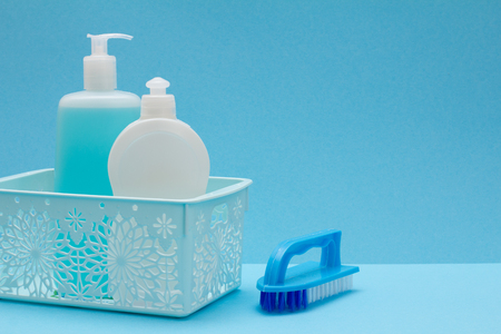 Plastic bottles of dishwashing liquid, glass and tile cleaner in basket and brush on blue background. Washing and cleaning concept.の写真素材