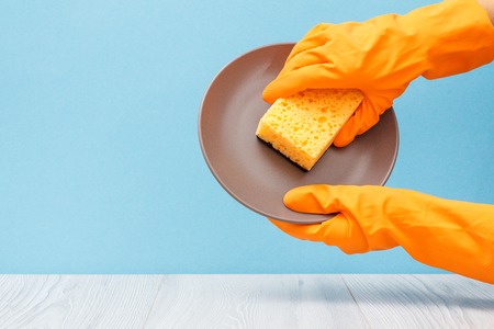 Women's hands in orange protective glove with plate and sponge on blue background. Washing and cleaning concept.の写真素材