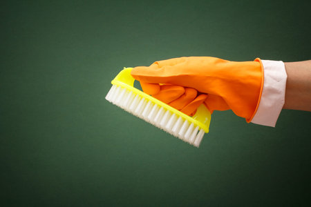 Woman's hand in orange protective glove with brush on green background. Washing and cleaning...