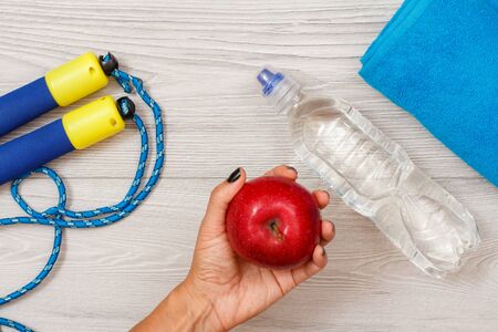 Female hand is holding a red apple in room or gym with jump rope, bottle of water and towel in the background. Tools for fitness. Top view.の写真素材
