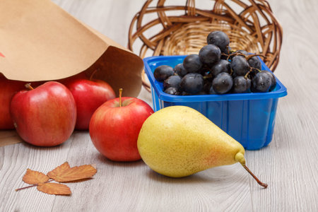 Ripe yellow pear, bunch of grapes in a box, wicker basket, red apples with paper bag and dry leaf on...