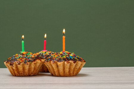 Three tasty birthday cupcakes with chocolate glaze and caramel, decorated with burning festive candles on green background with copy space. Happy birthday minimal concept.の写真素材