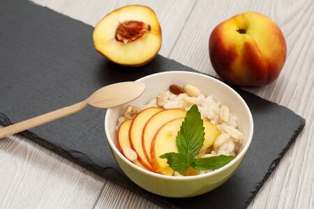 Porcelain bowl with sorghum porridge, cashew nuts, almond, whole fresh peach and a half one, wooden spoon on stone board.の写真素材
