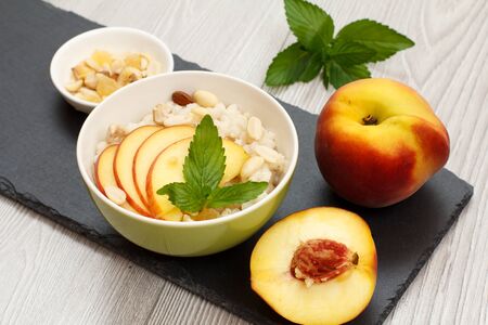 Porcelain bowl with sorghum porridge with cashew nuts and almond, whole fresh peach and a half one, small bowl with nuts on stone board.の写真素材