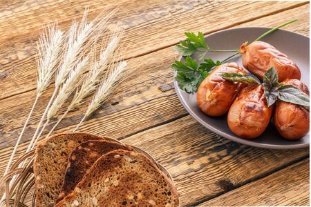 Toasts in wicker basket and grilled sausages with basil and parsley on plate. Top view with ears of wheat and wooden boards on the background..の写真素材