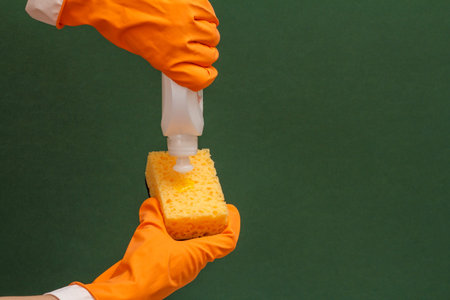 Women's hands in orange protective glove with bottle of dishwashing liquid and sponge on the green background. Washing and cleaning concept.の写真素材